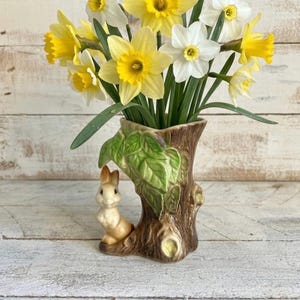 Può includere: Un bouquet di narcisi gialli e bianchi in un vaso di ceramica a forma di tronco d'albero con una statuina di coniglio. Il vaso presenta foglie verdi e un tronco marrone. I fiori hanno centri gialli e petali bianchi o gialli.