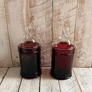 Peut inclure: Deux bocaux de pharmacie en verre avec des bouchons transparents et un contenu rouge foncé. Les bocaux sont cylindriques avec une base arrondie et un col étroit. Ils reposent sur une surface en bois peinte en blanc.