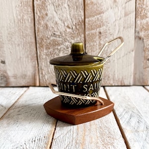 Peut inclure: Un pot à condiments en céramique vert olive foncé avec un couvercle et une cuillère en métal, présentant un motif géométrique texturé et le texte MINT SAUCE imprimé sur le côté. Le pot est maintenu par un cadre métallique sur une base en bois.