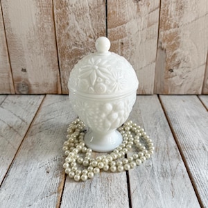 Pode incluir: Um pote de vidro leitoso branco com tampa, apresentando um padrão floral e de frutas em relevo no exterior. O recipiente é sustentado por uma base de pedestal e está rodeado por um colar de pérolas brancas artificiais para exibição.