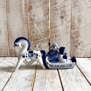 May include: A white ceramic figurine featuring a horse pulling a sled with two figures seated inside. The piece is decorated with blue painted details, including floral patterns on the sled and circular motifs on the horse's body and mane.