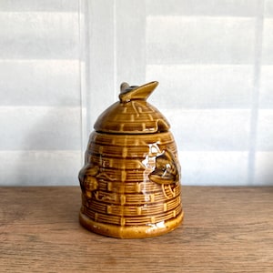May include: A brown ceramic honey pot with a lid, shaped like a beehive. The pot has a textured surface with bee and honeycomb details. The lid has a small handle. The honey pot is sitting on a wooden surface.