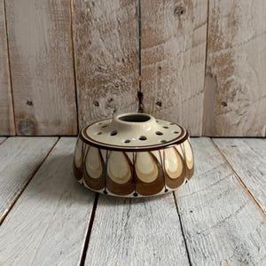 May include: A ceramic flower frog with a cream-coloured top and a brown and cream teardrop pattern around the sides. The flower frog has multiple holes for arranging flowers and is set on a white wooden surface.