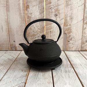 May include: A matte black cast iron teapot with a textured hobnail pattern on the exterior, featuring a curved handle, a small lid with a knob, and a matching circular trivet base. The teapot has a short spout and a rounded body shape.