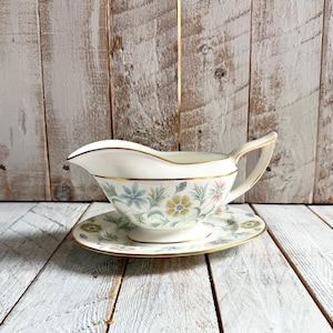 Könnte beinhalten: Eine weiße Saucieren aus Porzellan mit passender Untertasse, verziert mit einem zarten Blumenmuster in Blau-, Gelb- und Grüntönen. Das Set ist mit einem dünnen Goldrand an den Kanten der Sauciere und der Untertasse sowie einem geschwungenen Griff versehen.