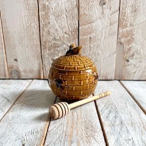 May include: A brown ceramic honey pot shaped like a beehive with a textured basket-weave pattern and a small bee figurine on the lid. A light-colored wooden honey dipper with a grooved head rests in front of the pot on a white wooden surface.