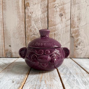 May include: A deep purple ceramic lidded jar featuring a rounded body with two side handles and a matching lid. The word BEETROOT is embossed on the lid. The jar has a glossy finish and decorative embossed swirl patterns on the front surface.