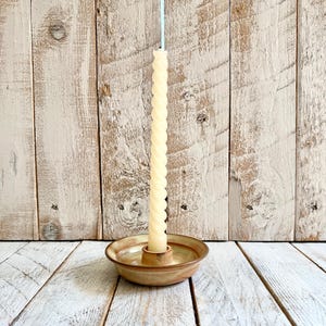May include: A cream-coloured, spiral-shaped candle stands in a small, round, ceramic candle holder. The candle holder has a light brown and tan colour scheme. The background is a light-coloured wooden wall and floor.