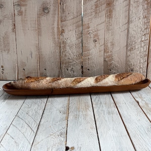 May include: A crusty baguette sits in a long, wooden serving dish. The bread has a golden-brown crust with visible scoring and a dusting of flour. The dish is a rich, dark brown, set against a rustic, white-washed wooden backdrop.