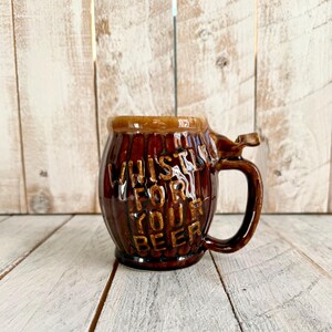 Può includere: Un boccale da birra in ceramica marrone con manico e le parole "Whistle for your beer" in rilievo sul davanti. Il boccale ha una forma a botte e una glassa marrone scuro.