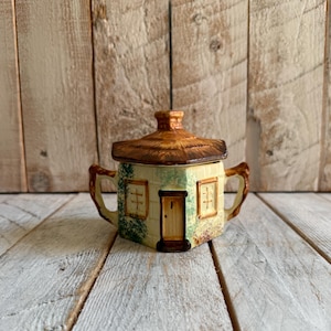 May include: A ceramic sugar bowl shaped like a small cottage with a removable thatched-style roof lid. The piece features cream-colored walls, brown trim, two side handles, and painted details including a central door, windows, and greenery.