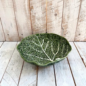 May include: A dark green ceramic bowl shaped like a cabbage leaf with a glossy finish. The bowl features a detailed, embossed texture mimicking the veins and crinkled surface of a real leaf, with a scalloped edge and a prominent central stem.