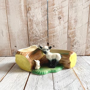 Könnte beinhalten: Ein Keramik-Pflanzgefäß in Form eines Baumstamms mit einem Lamm und einem kleinen Hasen auf einer grünen Basis. Der Baumstamm hat gelbe und braune Akzente mit einer ausgehöhlten Mitte zum Bepflanzen. Das Lamm hat schwarz-weiße Markierungen.