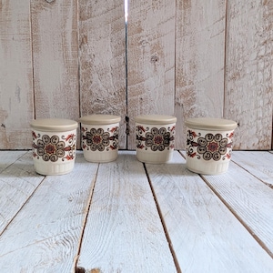 Puede incluir: Cuatro botes de cerámica vintage con tapas. Los botes son de color blanquecino con un patrón repetido de diseños florales marrones, rojos y dorados. Las tapas son de color blanquecino liso. Los botes están dispuestos en fila sobre una superficie de madera blanca.