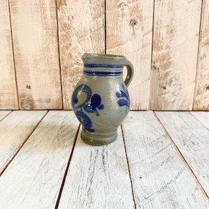 Op de afbeelding: Kleine, grijze keramische kruik met een handvat en een afgeronde body. De kruik heeft blauwe bloemmotieven en horizontale blauwe strepen rond de hals. De achtergrond is een lichtgekleurd houten oppervlak.