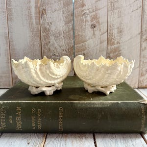 Puede incluir: Dos cuencos de cerámica en forma de concha, de color blanquecino. Los cuencos tienen un borde con volantes y se asientan sobre una pequeña base decorativa. Se exhiben sobre un libro verde vintage con el título "Pottery and Porcelain".