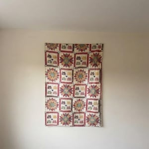 May include: A rectangular quilt featuring a repeating pattern of multi-coloured star blocks alternating with blocks displaying small house illustrations in red, green, yellow, and black, all set against a cream background with red borders.