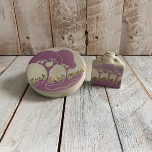 May include: A ceramic set featuring a round lidded container and a rectangular container. Both pieces are off-white with a purple tree design and colorful accents. The set is placed on a white wooden surface.