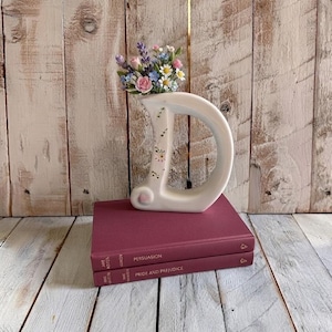Può includere: Un vaso in ceramica bianca a forma di lettera D, decorato con piccoli fiori dipinti e riempito con un bouquet di lavanda, rose rosa e margherite bianche artificiali, appoggiato su due libri impilati color bordeaux intitolati Persuasion e Pride and Prejudice.
