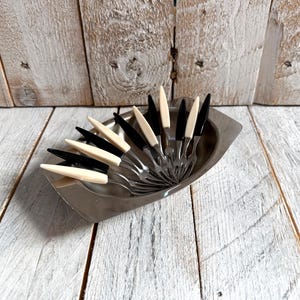 May include: A collection of vintage appetizer forks with black and cream-coloured handles, presented in a silver serving dish. The forks are arranged on a weathered, white-painted wooden surface, creating a rustic aesthetic. The forks are approximately 13 cm long.