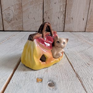 Puede incluir: Una figurita de cerámica de un bolso amarillo y rojo con un asa marrón. Un pequeño gato blanco asoma por la parte superior del bolso.