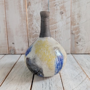 May include: A small, round ceramic vase with a narrow neck. The vase is painted in shades of white, yellow, blue, and grey.