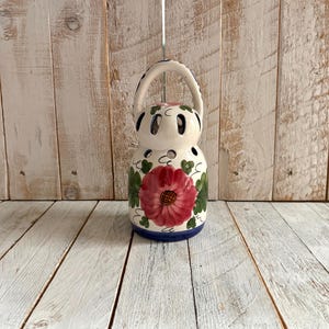 May include: A white ceramic candle holder with a handle, decorated with a red and pink floral design and green leaves. The holder has a blue base and small holes for light. The handle is white with blue accents. A decorative item.