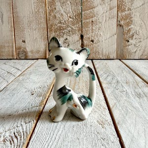 May include: A vintage ceramic cat ornament. The cat is white with black and teal detailing on the ears, tail, and back. It has a red mouth and black eyes. The figurine is in a seated pose.