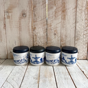 Może przedstawiać: Cztery białe ceramiczne słoiki z ciemnoniebieskimi pokrywkami. Każdy słoik ma niebiesko-białą ilustrację holenderskiego wiatraka i sceny wioski. Słoiki są ułożone w rzędzie na tle rustykalnego drewna.