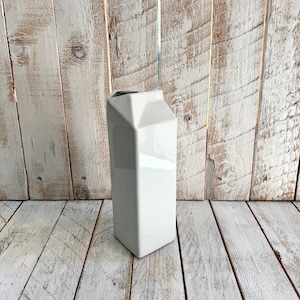 Può includere: Un vaso di ceramica bianca a forma di cartone del latte. Il vaso ha una base rettangolare e una parte superiore piegata, con una finitura lucida. Lo sfondo è un muro e un pavimento in legno rustico.