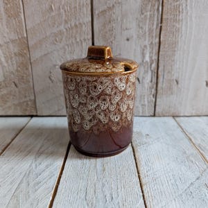 Puede incluir: Un tarro de cerámica con tapa, con un patrón moteado marrón y crema. El tarro tiene una base marrón oscuro que se desvanece en el diseño moteado color crema. La tapa tiene una pequeña asa y una ranura para una cuchara. El tarro es probablemente para almacenar alimentos.