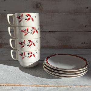 May include: A stack of four white ceramic teacups with a red and black leaf design. The cups are stacked on a white wooden surface. There are four matching saucers with a red and gold trim stacked next to the cups.