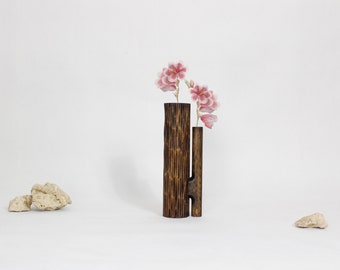 Puede incluir: Un jarrón de madera con un diseño único, que presenta dos cilindros conectados. El jarrón es de color marrón oscuro y tiene una superficie texturizada. Flores rosas están dispuestas en el jarrón.