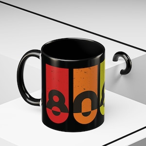 May include: A black ceramic mug featuring a graphic design with the number 80 in a distressed, retro-inspired font. The design consists of vertical rectangular panels in red, orange, and yellow-green, each containing a portion of the black number 80.
