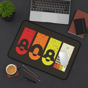 Può includere: Tappetino per mouse rettangolare nero con il numero "808" in rosso, arancione, giallo e bianco. Il tappetino ha un aspetto vintage e usurato. Un mouse per computer bianco è posizionato sul tappetino, con un laptop e un taccuino sullo sfondo.