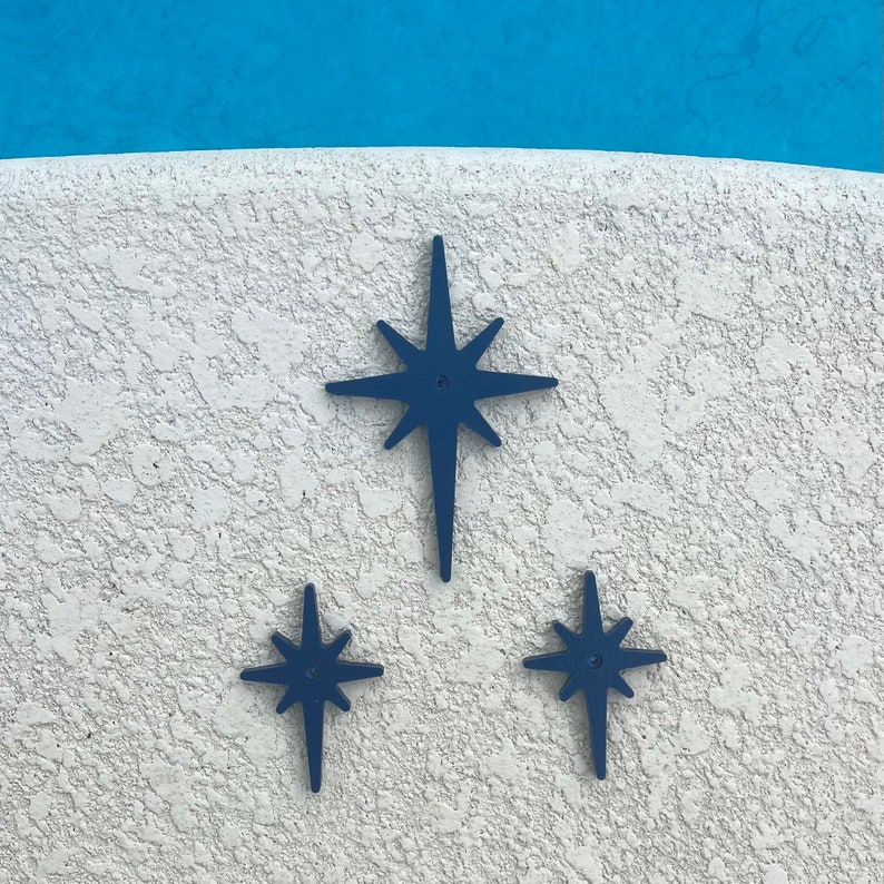 May include: Three blue starburst shapes are mounted on a white textured wall. The largest starburst is in the center and the two smaller starbursts are on either side of the larger starburst.