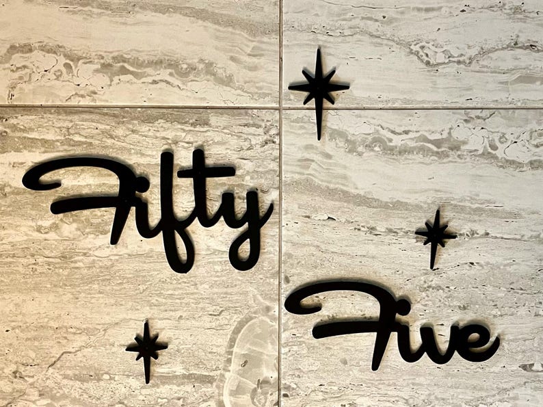 May include: Black wooden cutout of the words "Fifty Five" with three black star shapes on a light brown marble background.