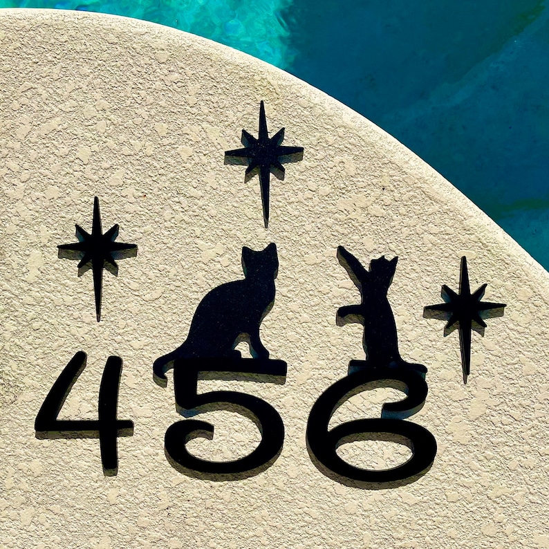 May include: A white ceramic house number plaque with black silhouettes of two cats and three stars. The numbers 456 are displayed in black.