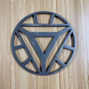 May include: A silver metal wall decor in the shape of the Iron Man symbol. The symbol is a circle with a triangle inside, and the circle is surrounded by a series of smaller circles.