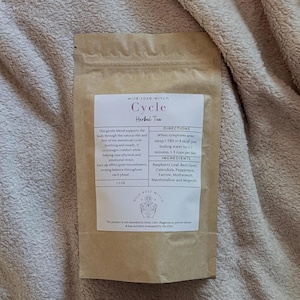 May include: A brown paper packet of "Cycle Herbal Tea" from Wild Rose Witch. The label details ingredients such as raspberry leaf and peppermint. The text describes the tea's benefits for the menstrual cycle and brewing instructions.