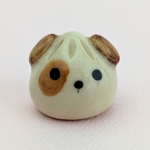 May include: A small ceramic figurine of a dog with brown ears and spots. The dog has a white body and black eyes.