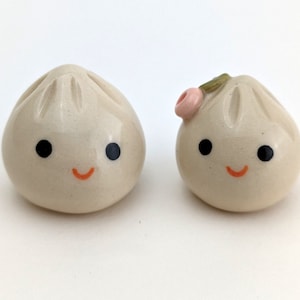 Op de afbeelding: Twee kleine keramische beeldjes in de vorm van gestoomde broodjes met blije gezichten. Het broodje rechts heeft een roze bloemetje erop.