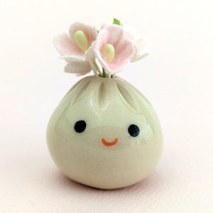 Peut inclure: Un petit vase en céramique en forme de boulette avec un visage souriant et des fleurs roses sur le dessus.