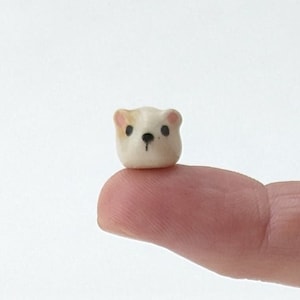 May include: A small white ceramic bear figurine with black eyes and a pink nose. The bear is sitting on a finger.