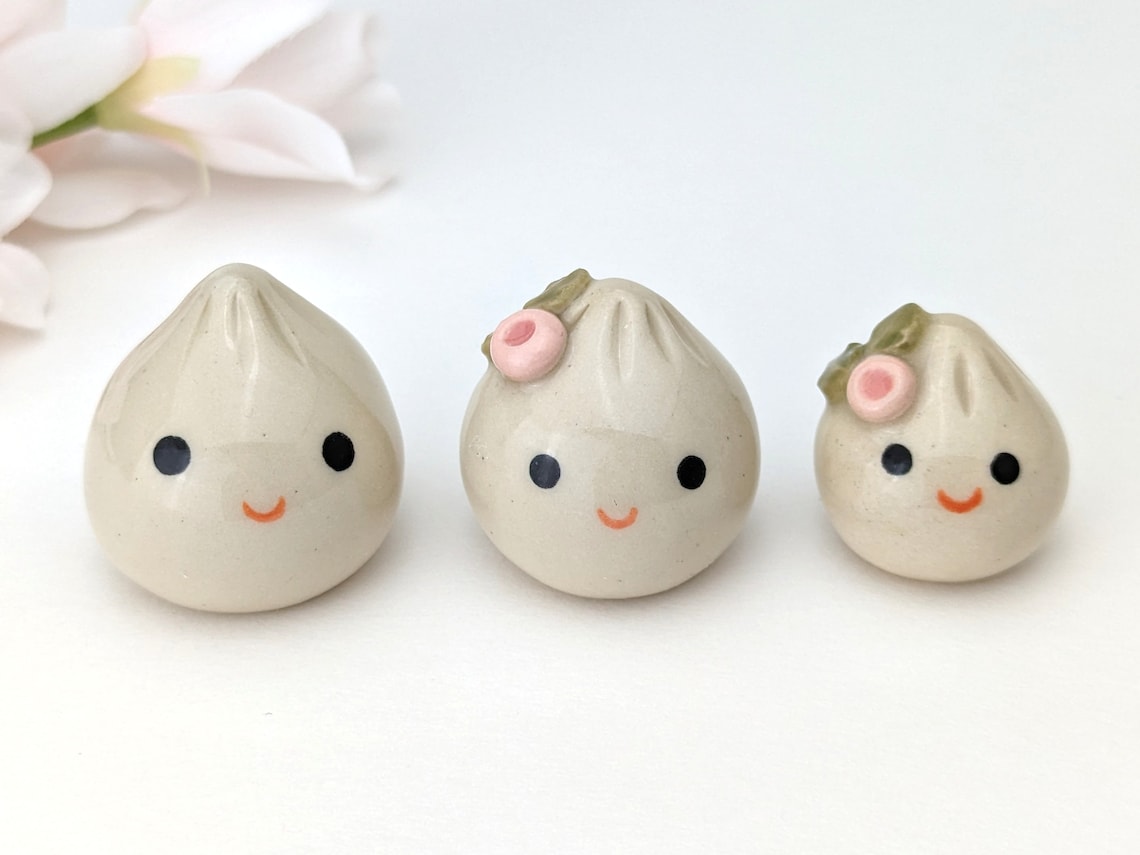 May include: Three ceramic figurines shaped like steamed buns with happy faces. The two larger buns have pink flowers on top and the smallest bun has a single pink flower on top.