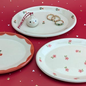 May include: Three decorative ceramic oval plates with floral and fruit designs. One plate has a cherry design, another has pink flowers, and the third has a dumpling ornament and rings. The plates are on a red surface.