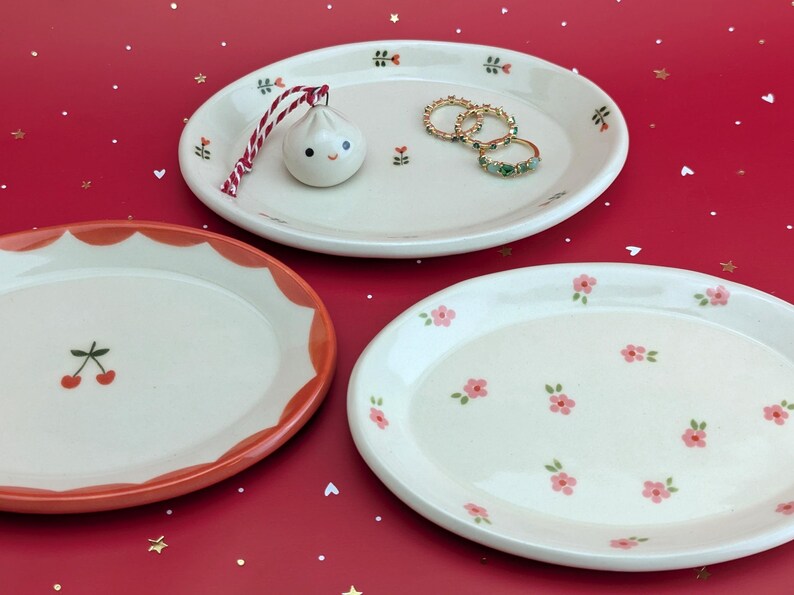 May include: Three decorative ceramic oval plates with floral and fruit designs. One plate has a cherry design, another has pink flowers, and the third has a dumpling ornament and rings. The plates are on a red surface.