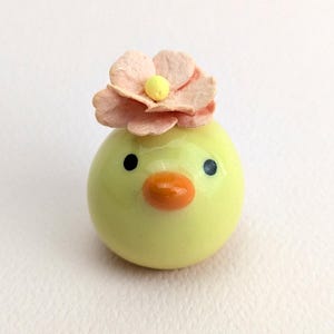 Könnte beinhalten: Ein kleines, gelbes Keramikküken mit einer rosa Blume auf dem Kopf. Das Küken hat schwarze Augen und einen orangefarbenen Schnabel.