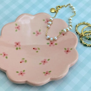 May include: A pink, scalloped-edge ceramic dish with hand-painted pink flowers and green leaves. A gold chain bracelet with white pearls and three gold rings with green stones rest on the dish. The background is a light blue checkered pattern.