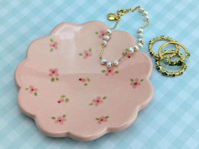 May include: A pink, scalloped-edge ceramic dish with hand-painted pink flowers and green leaves. A gold chain bracelet with white pearls and three gold rings with green stones rest on the dish. The background is a light blue checkered pattern.